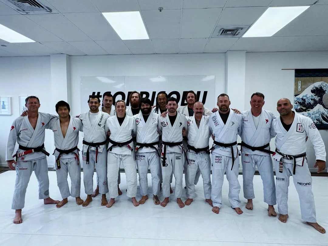 Twelve black belts in white gis standing arm in arm on the mats in front of the Sorriso BJJ wall
