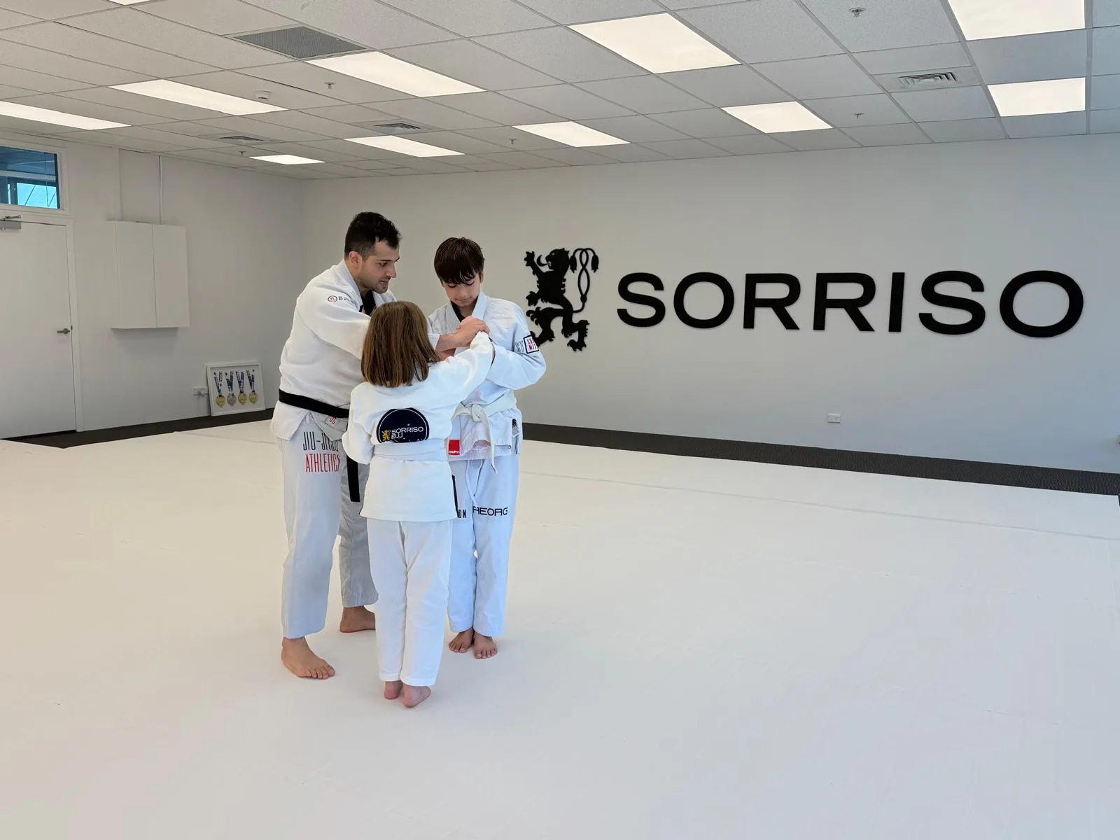 A black belt coach guides two children through a grip drill at a kids jiu-jitsu class in Botany Town Centre